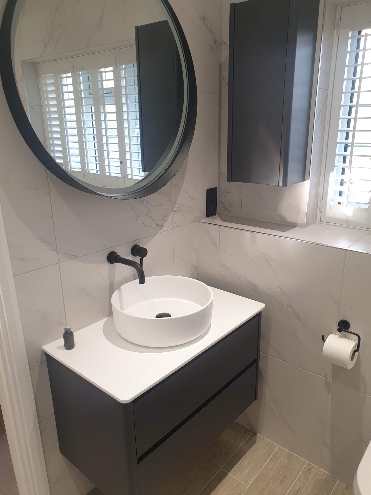 Marble-effect walls with a round mirror and matt black wall-mounted taps over a countertop basin, on a charcoal wall-hung vanity.