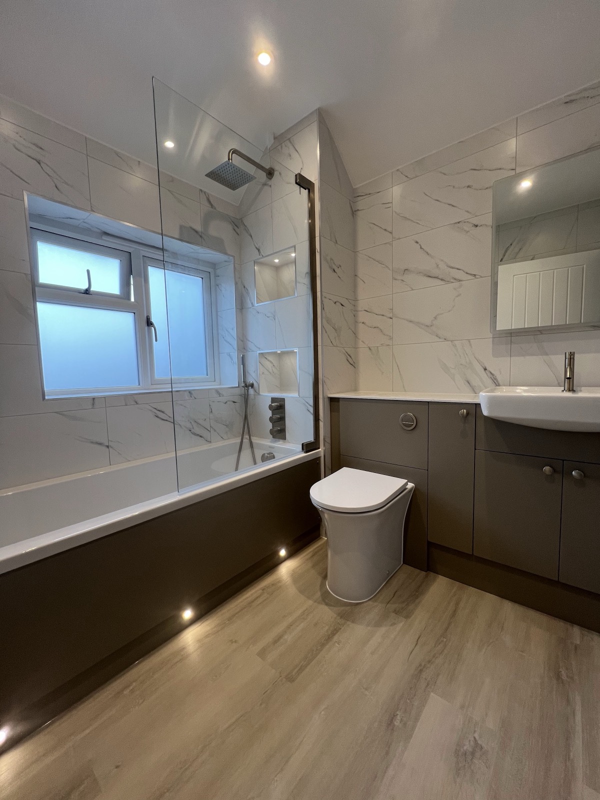 A bath-and-shower room in a sloping-ceiling space: marble-effect walls, plank-effect floor, a bath panel picked out in dark grey and plinth lighting set under it.