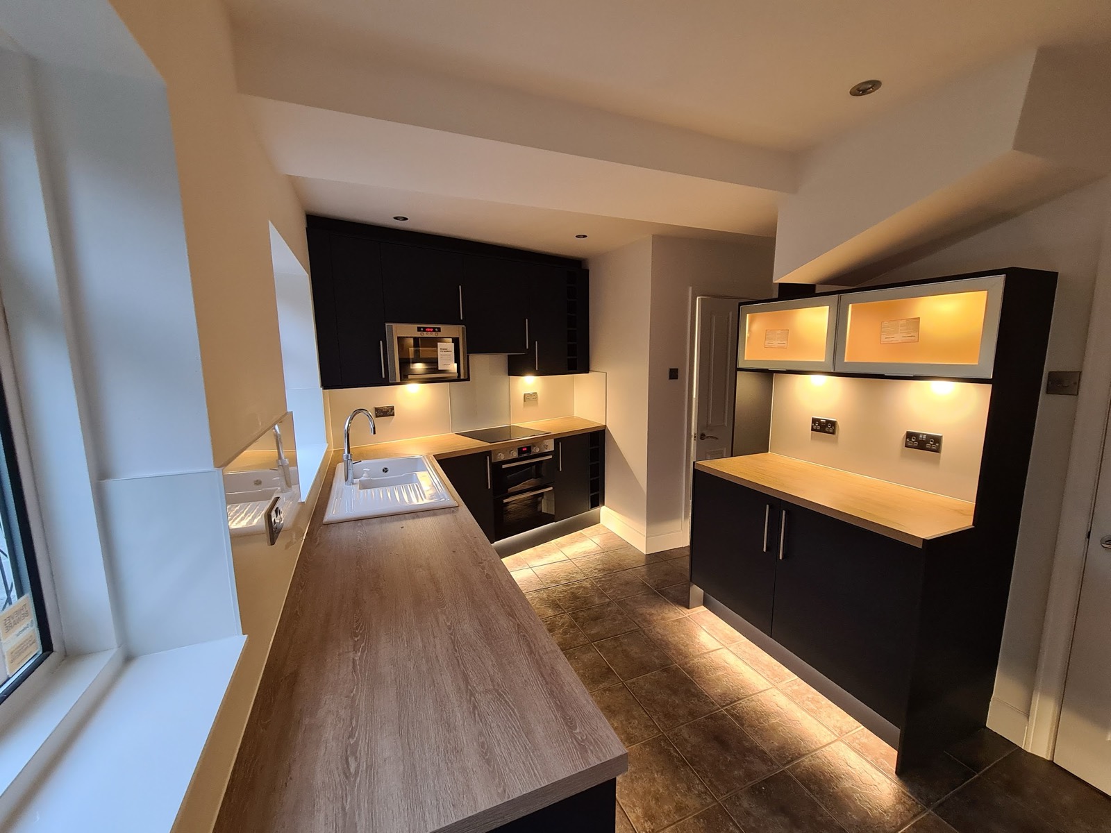 A galley kitchen fitted with dark units and oak-effect worktops, with under-cabinet and plinth lighting run in and an eye-level oven built into the run.