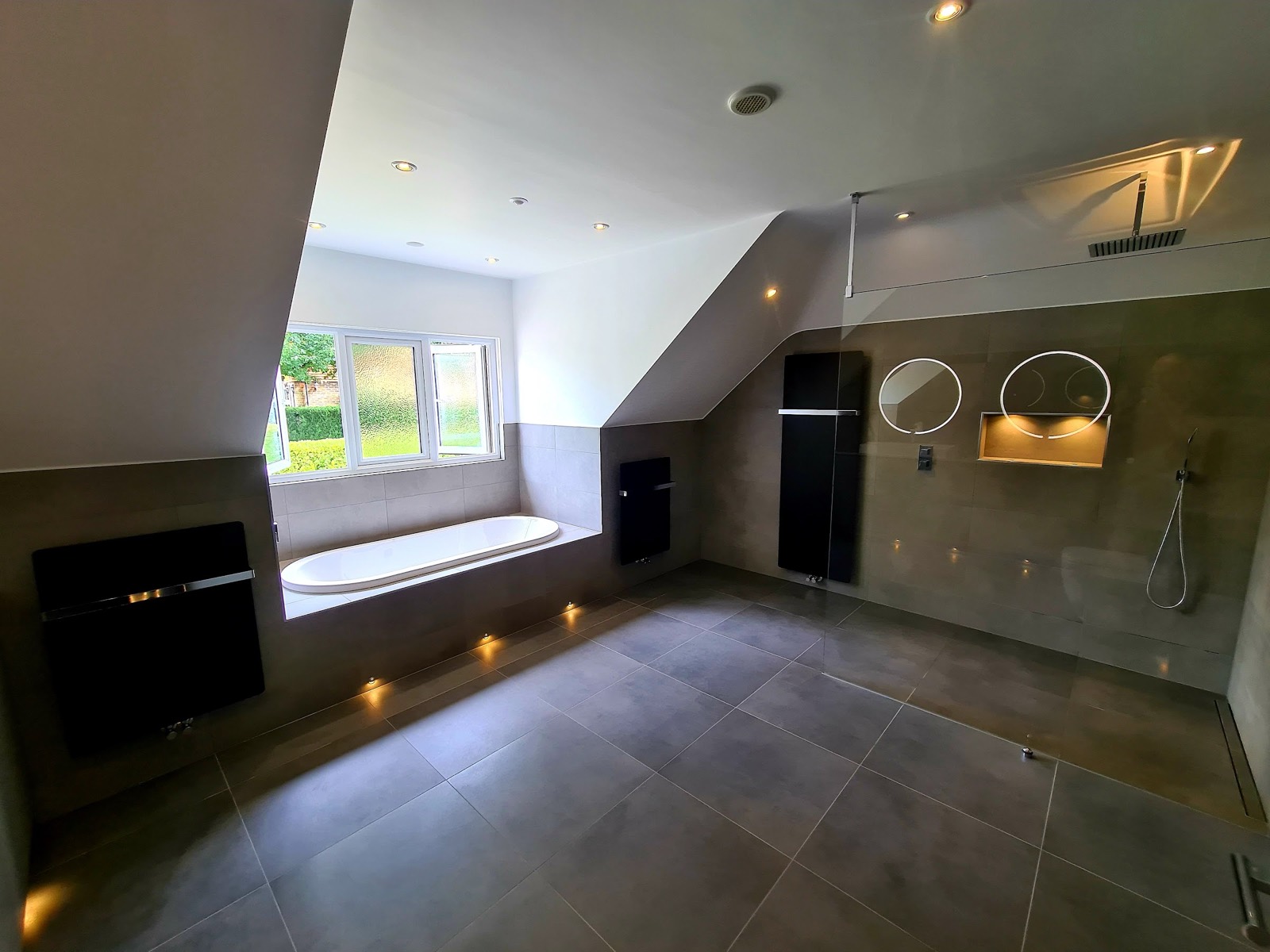A large room under a sloping ceiling: inset bath, big-format grey porcelain on walls and floor, black vertical radiators and floor-level lighting on a timer.