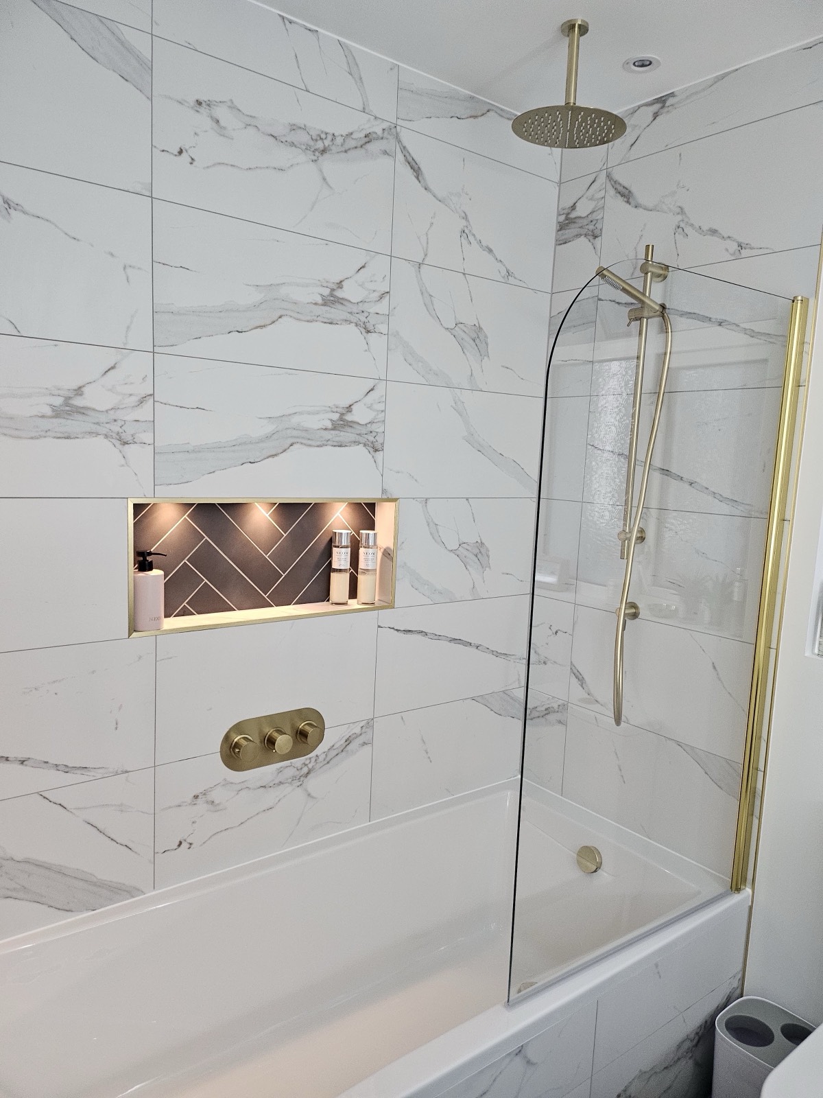Large-format marble-effect porcelain taken full height, with a herringbone-tiled recess set into the shower wall and a brushed brass rainfall shower over the bath.