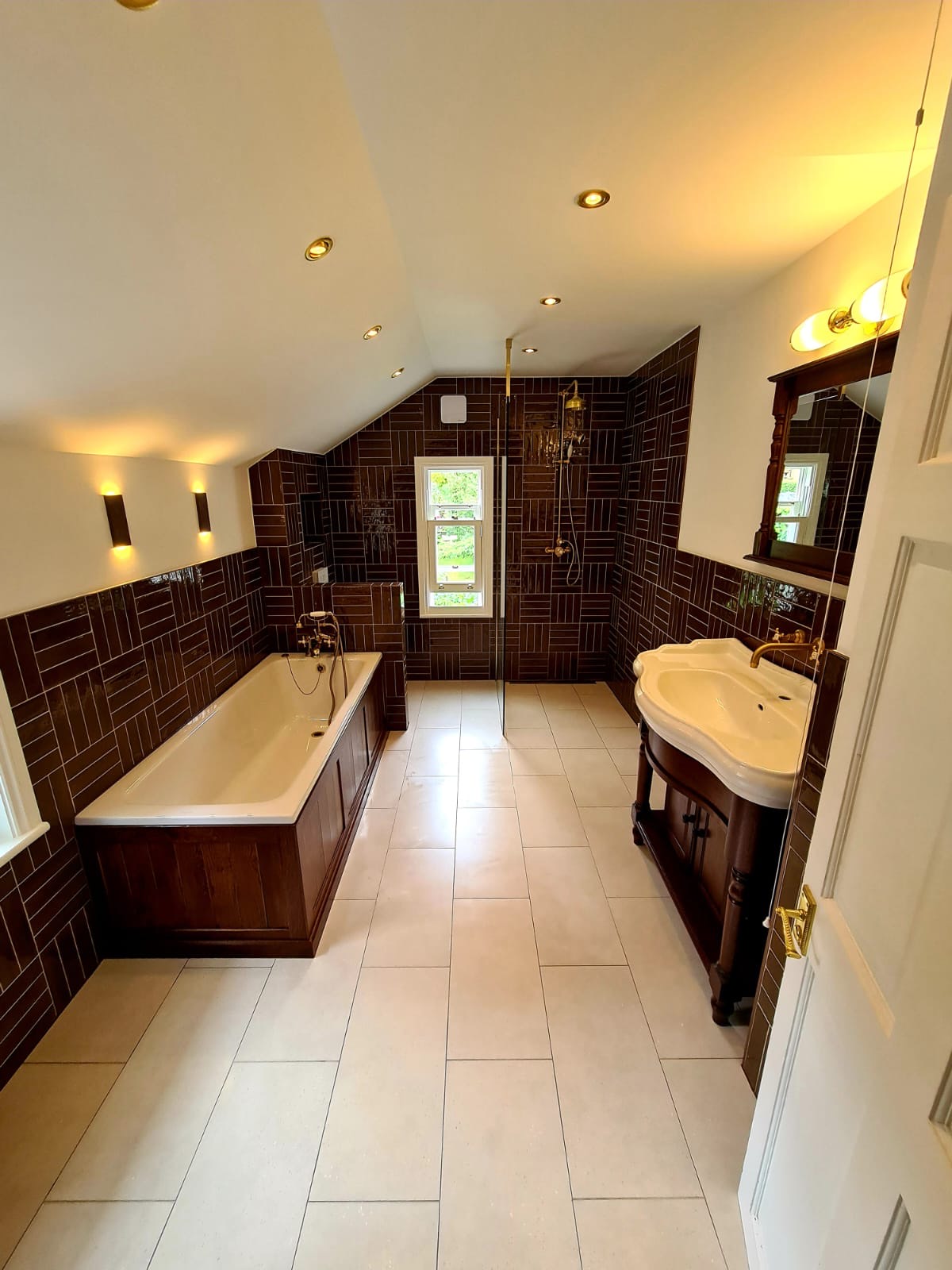 Traditional basin and panelled bath against a brown brick-bond tiled wall, with a walk-in shower and brass fittings — set out to suit the pitch of the ceiling.