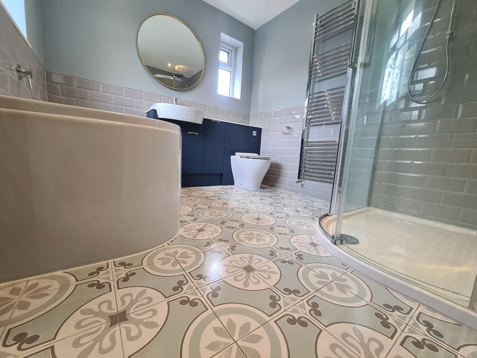 Patterned encaustic-style floor tiles set out so the pattern runs square to the room, with pale metro-tiled walls, a navy vanity and a quadrant shower enclosure.