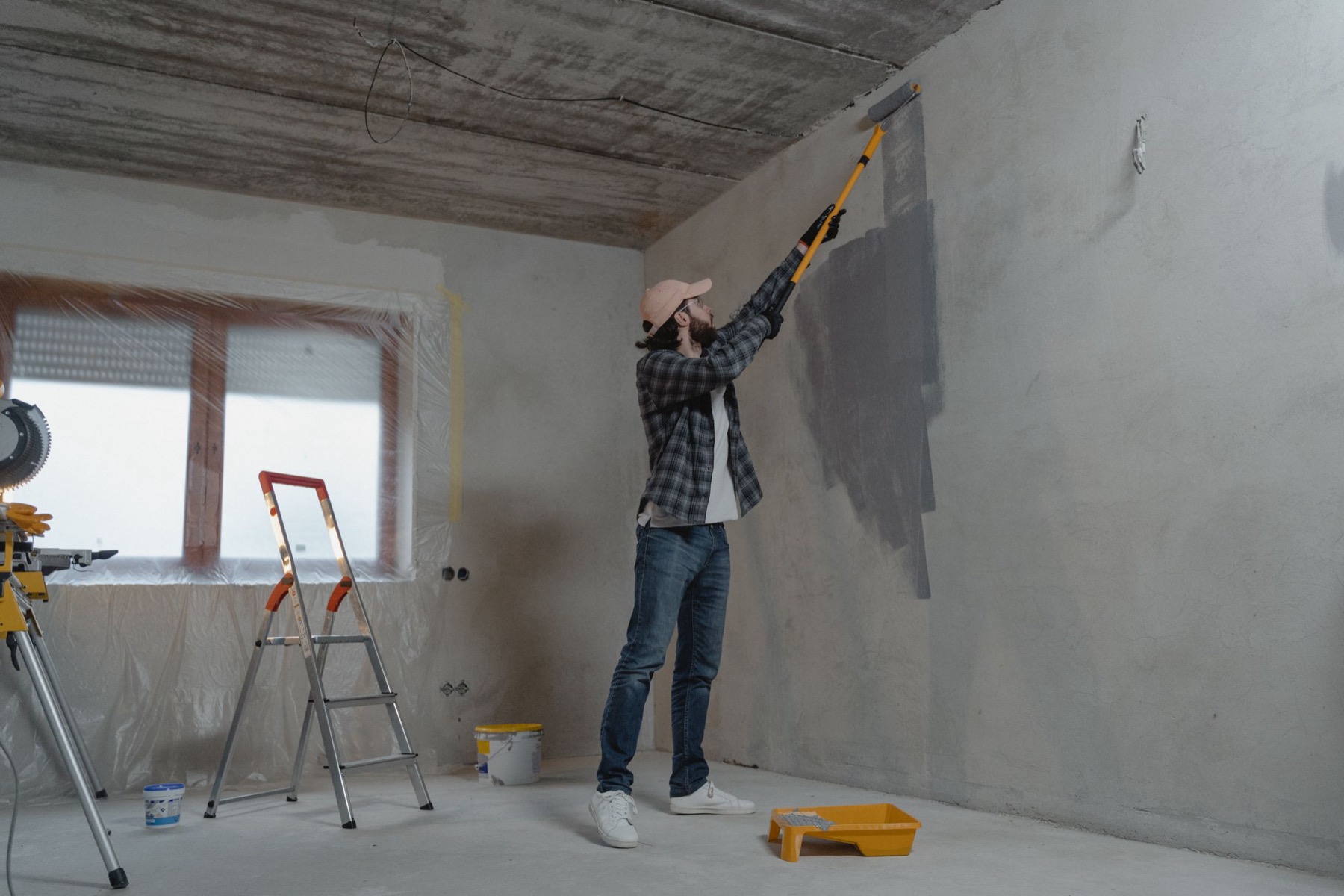 A tradesman skimming a bare wall during a refit, with a stepladder and paint tray on the protected floor